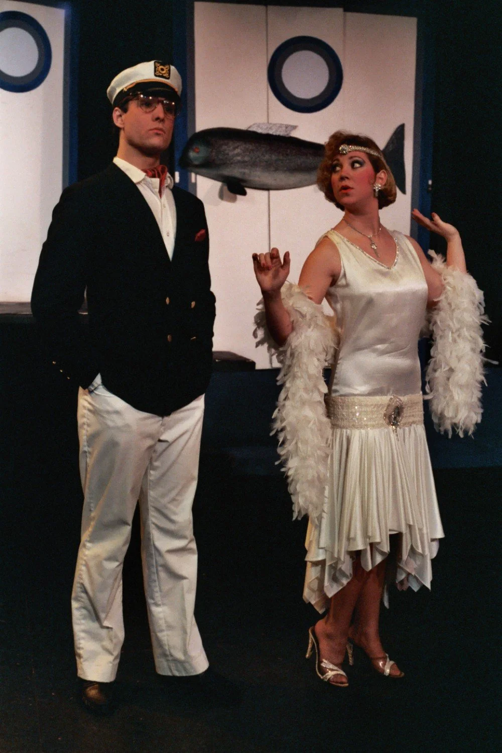 Two people in 1920s-inspired costumes, a man in a captain's outfit and a woman in a flapper dress, posing on stage.