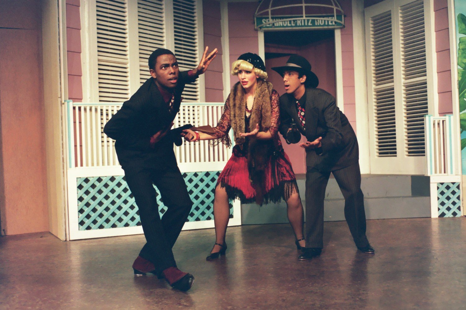 Three performers in 1920s costumes on stage, acting in a theatrical production in front of a hotel-themed backdrop.