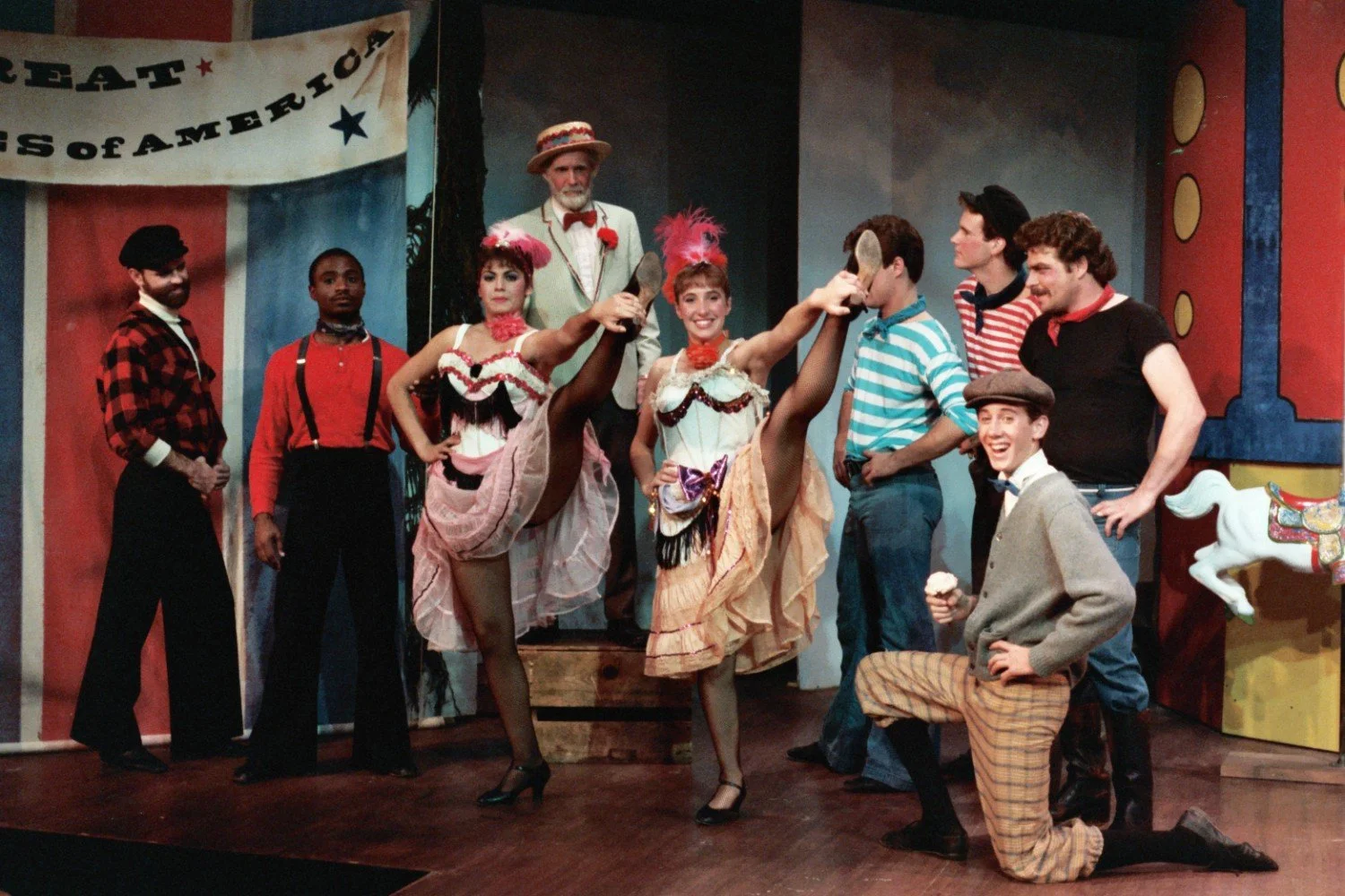 A group of performers in circus-themed costumes, including can-can dancers, posing on stage with a banner reading 'GREAT AMUSEMENTS of AMERICA'. The scene includes a background with carousel elements.