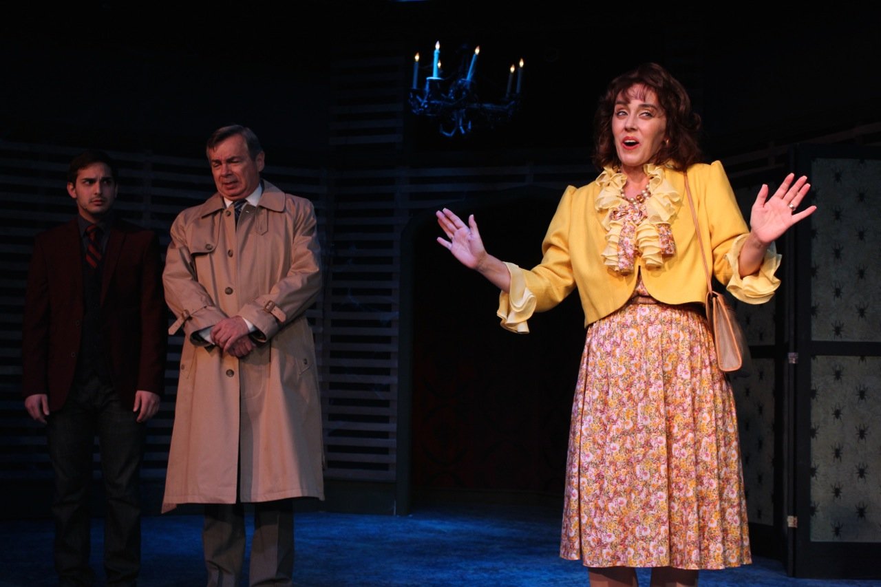 Three actors performing a scene on stage, with one woman in a yellow outfit and two men in suits, one wearing a trench coat, under dim lighting.