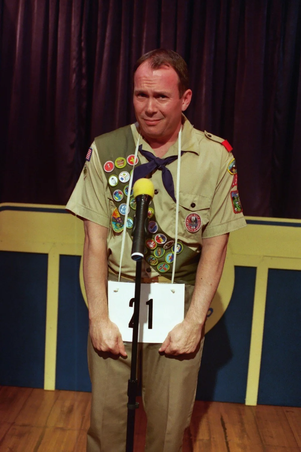 Man in a scout uniform standing on stage with a number 21 placard, in front of a microphone.