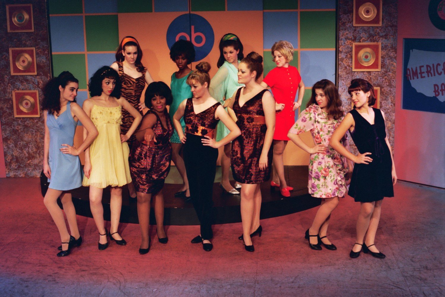 Group of women in 1960s-style dresses posing in a colorful room with retro decor.