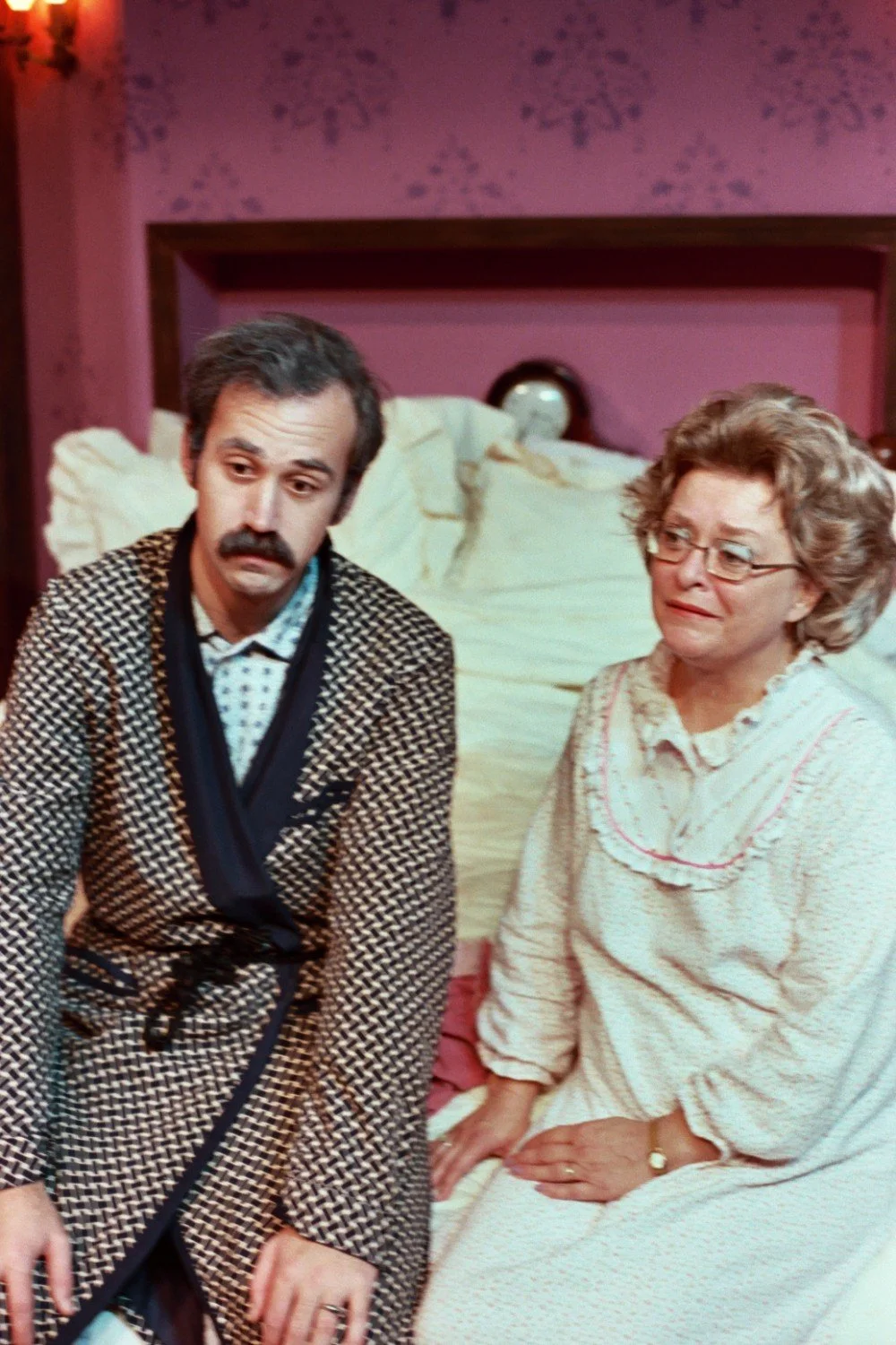 Two people sitting on a bed in a room with pink walls. The person on the left is wearing a patterned robe with a mustache, and the person on the right is wearing glasses and a nightgown.