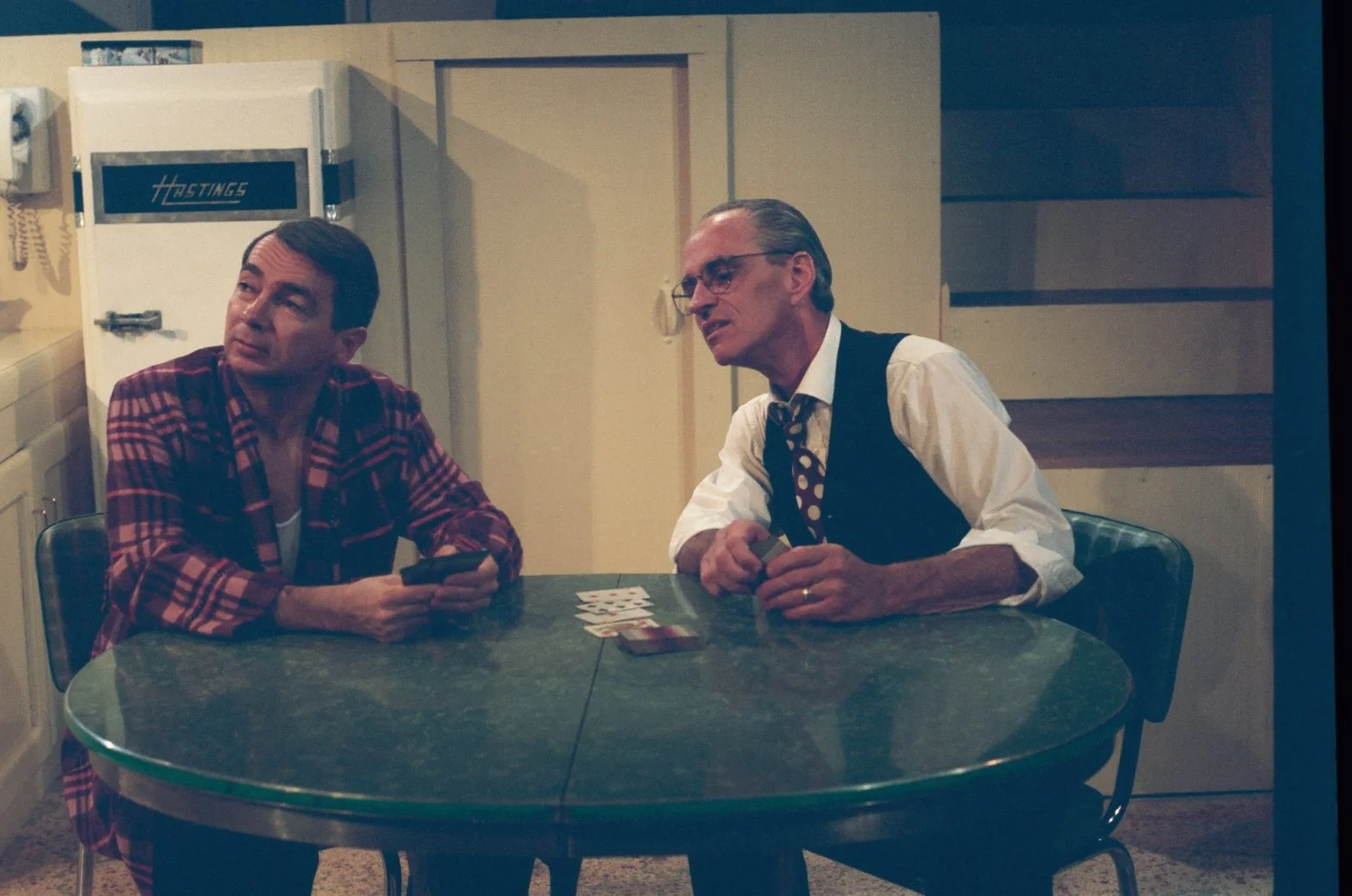 Two men sitting at a vintage kitchen table playing cards, with a white "Hastings" refrigerator in the background. One man is wearing a plaid robe, the other a dress shirt and tie with a vest. They appear engaged in conversation.