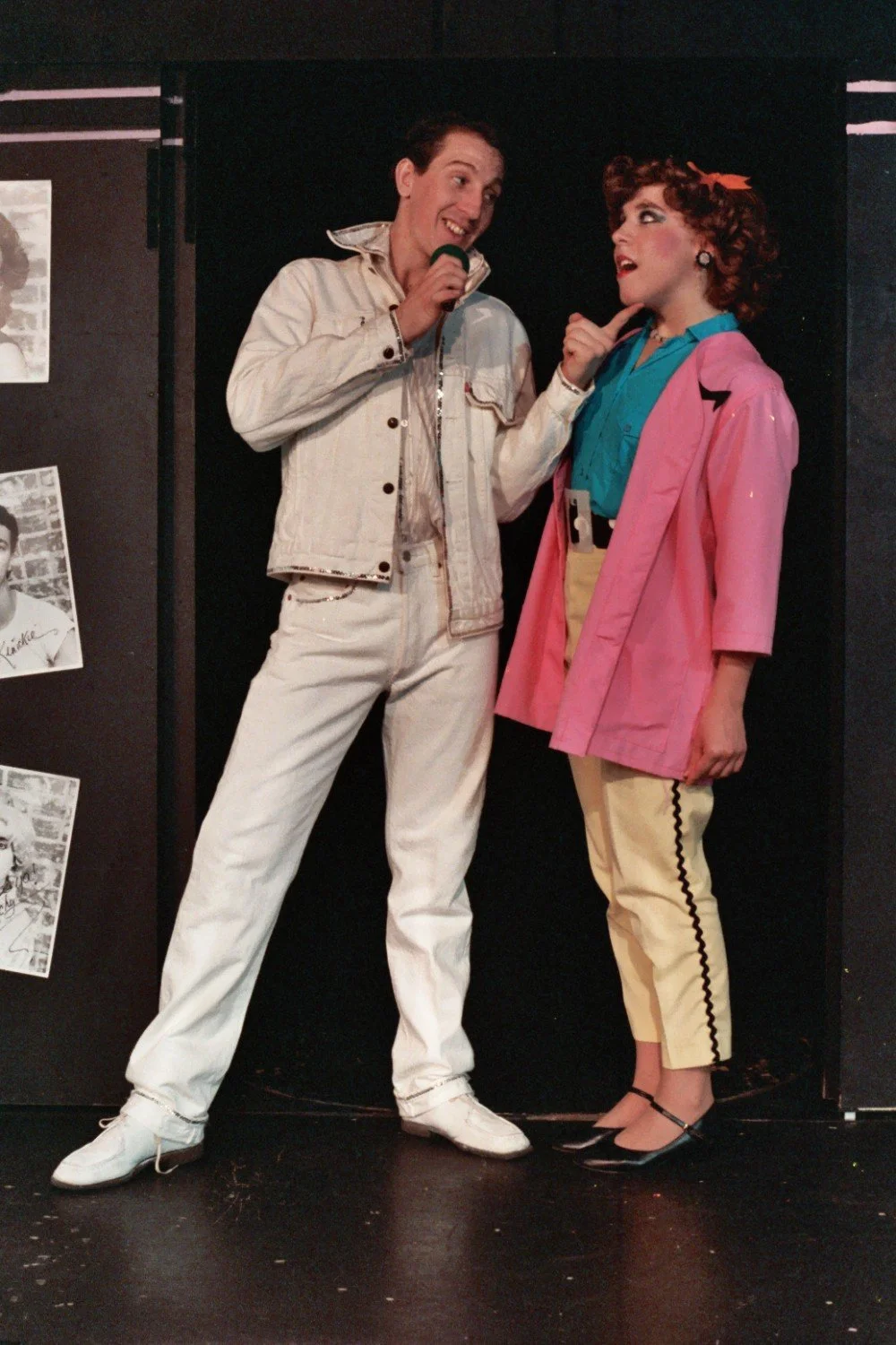 Two people on stage performing a musical scene, one in a white denim outfit singing into a microphone, the other wearing a pink jacket and colorful attire.