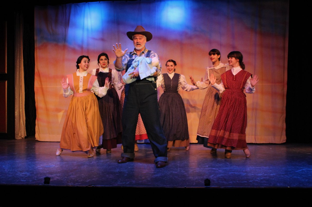 Stage performance featuring a man in a cowboy hat and five women in period costumes dancing.