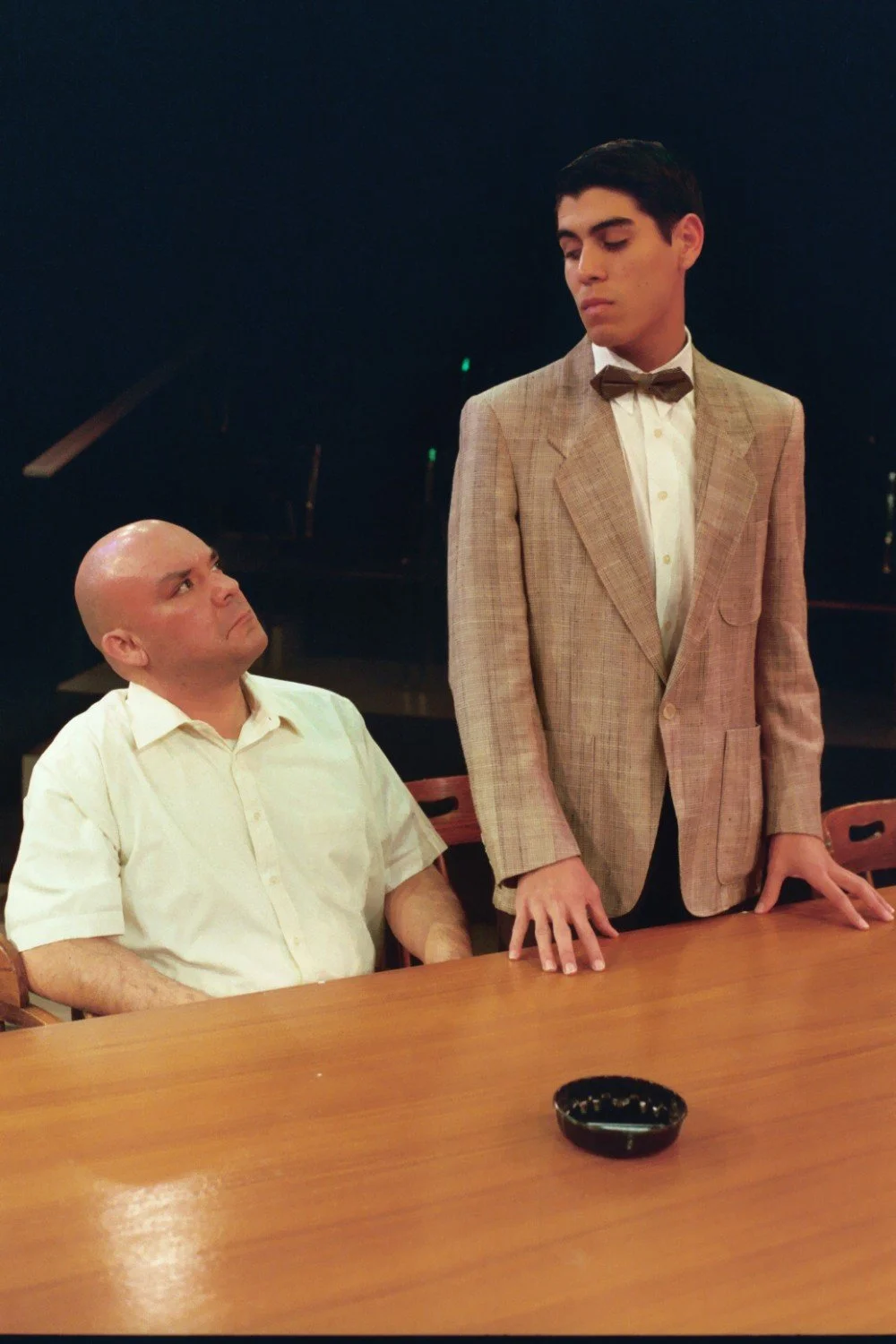 Two men in a dimly lit room, one standing in a suit with a bow tie, the other seated in a white shirt, positioned around a wooden table with an ashtray.