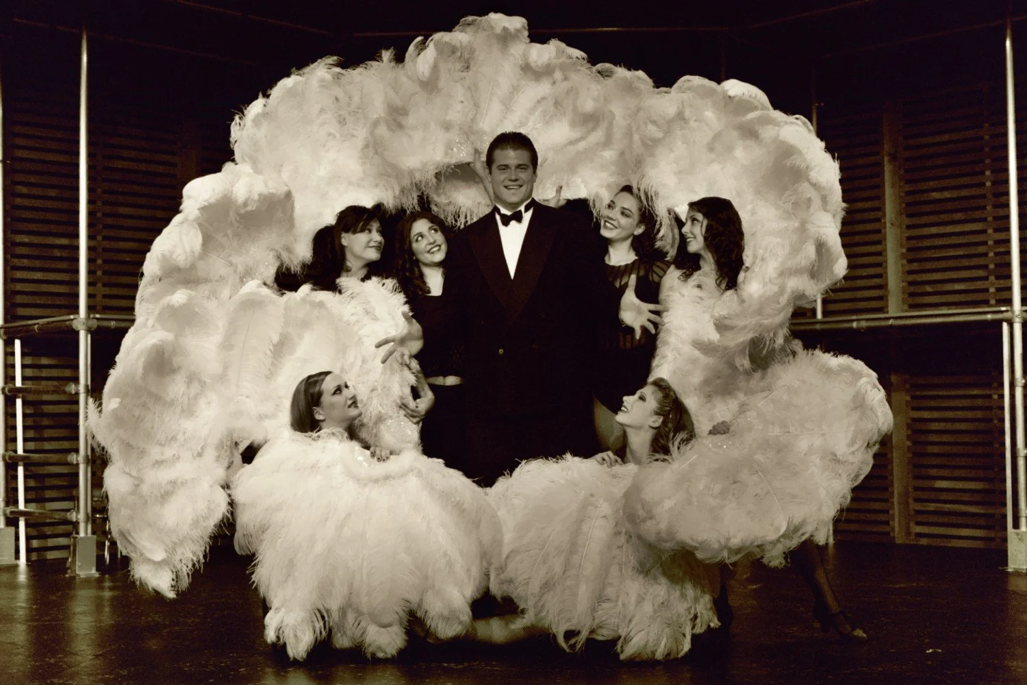 A group of dancers in white feathered costumes surrounding a man in a tuxedo on a stage.