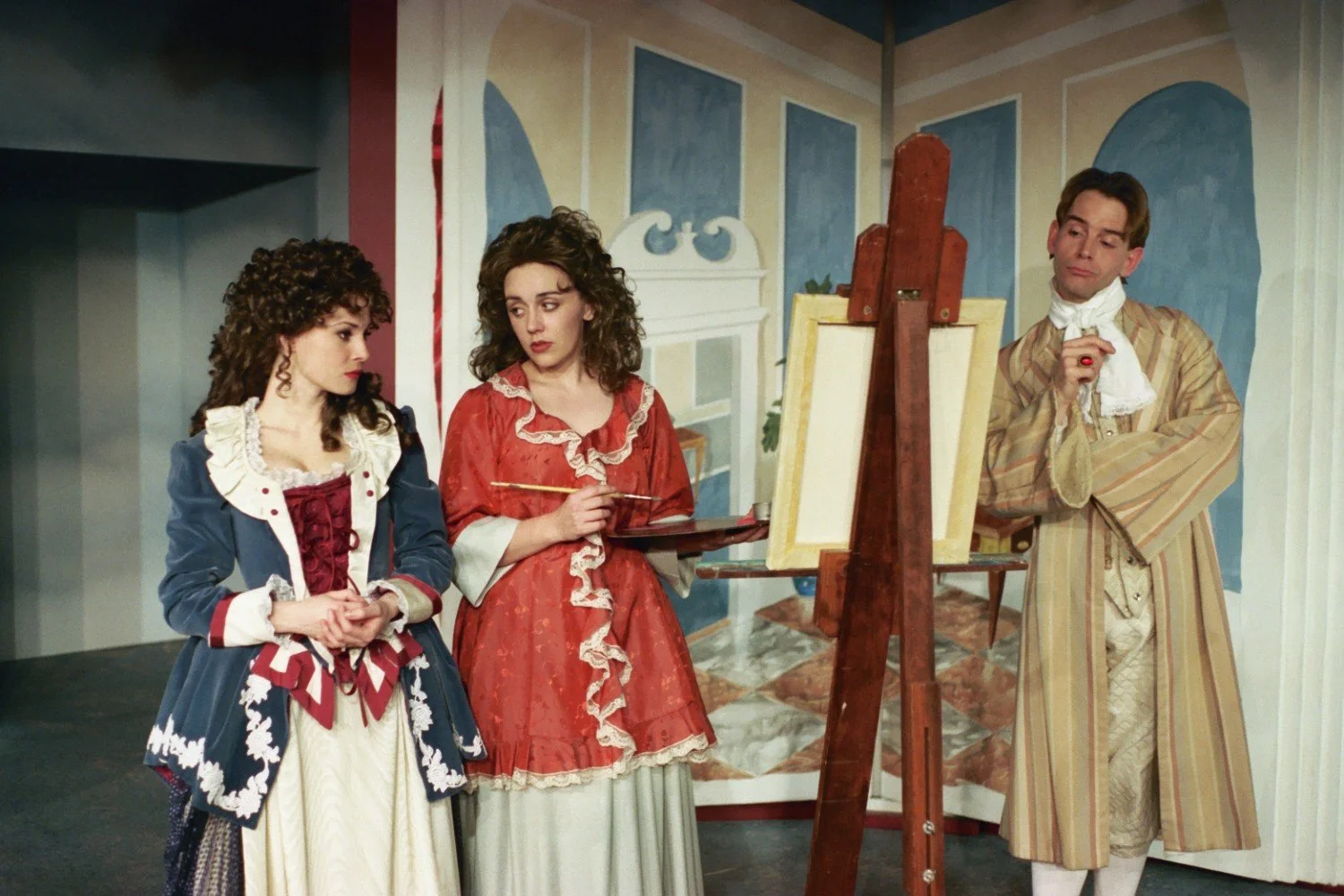 People in vintage costumes, likely actors in a play, around an easel in an artistic setting.