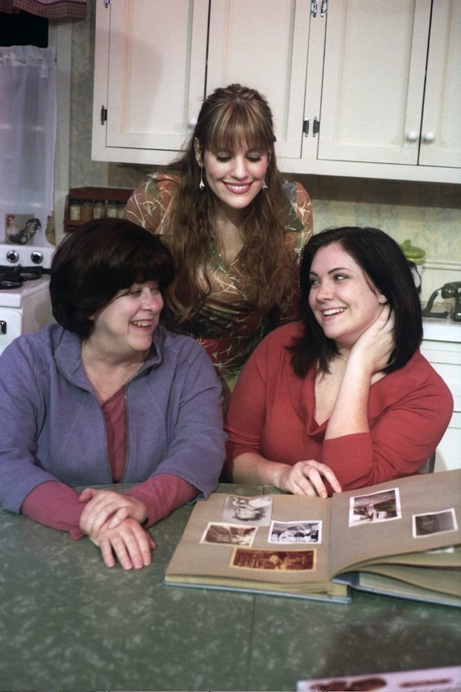 Three women sitting at a kitchen table, smiling and looking at a photo album.