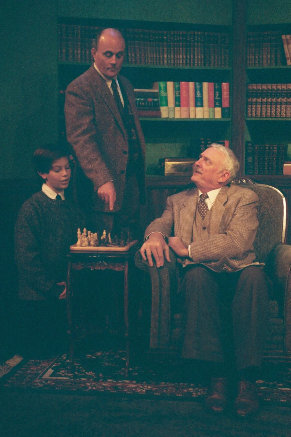 Three people in a study playing chess, surrounded by bookshelves.