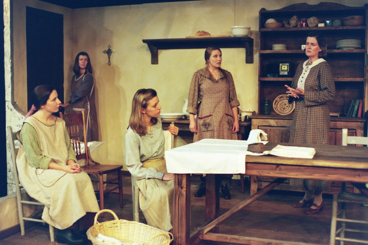 Theater scene with five women in period clothing, standing and seated around a wooden dining table with cloth. Shelves with pottery and a crucifix decorate the background.