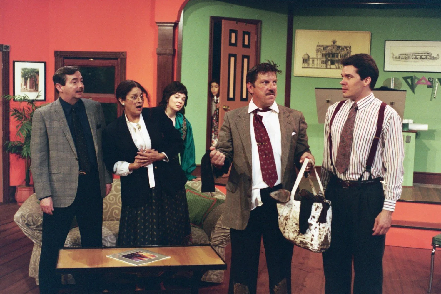 A group of people in a room, appearing involved in a dramatic scene. One man holds a dirty bag, while others look surprised. The room has colorful walls, framed art, and a potted plant.