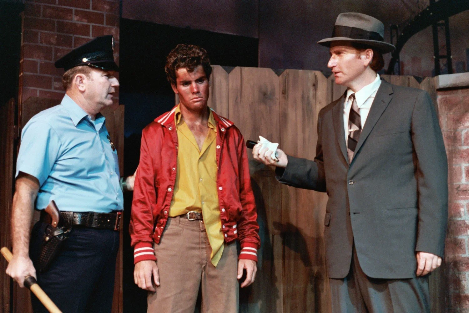 A scene with a police officer, a young man in a red jacket, and a man in a suit and hat, standing in front of a fence.