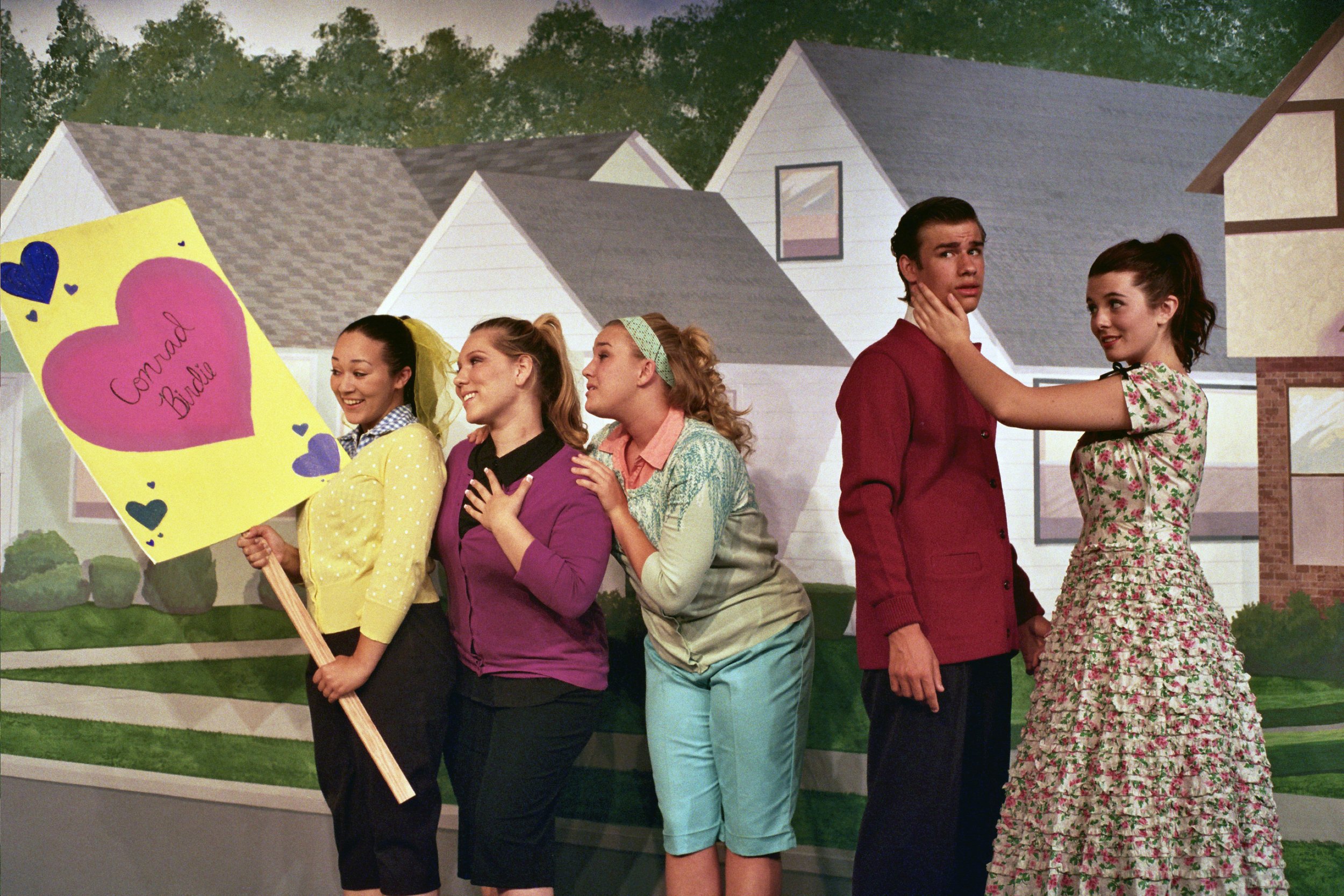 Group of actors on stage performing a scene with a backdrop of suburban houses. A woman holds a sign saying "Conrad Birdie," and others appear engaged in an animated, theatrical moment.