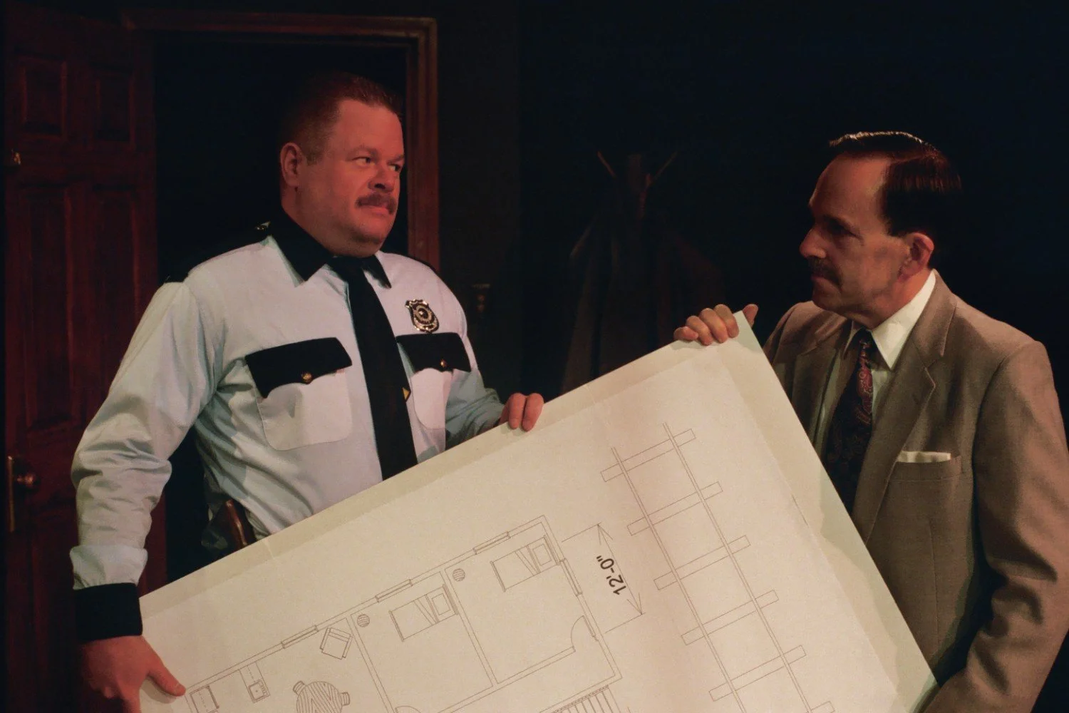 A police officer in uniform and a man in a suit holding a large blueprint. The blueprint features architectural floor plans. They appear to be discussing the document in a dimly lit room.