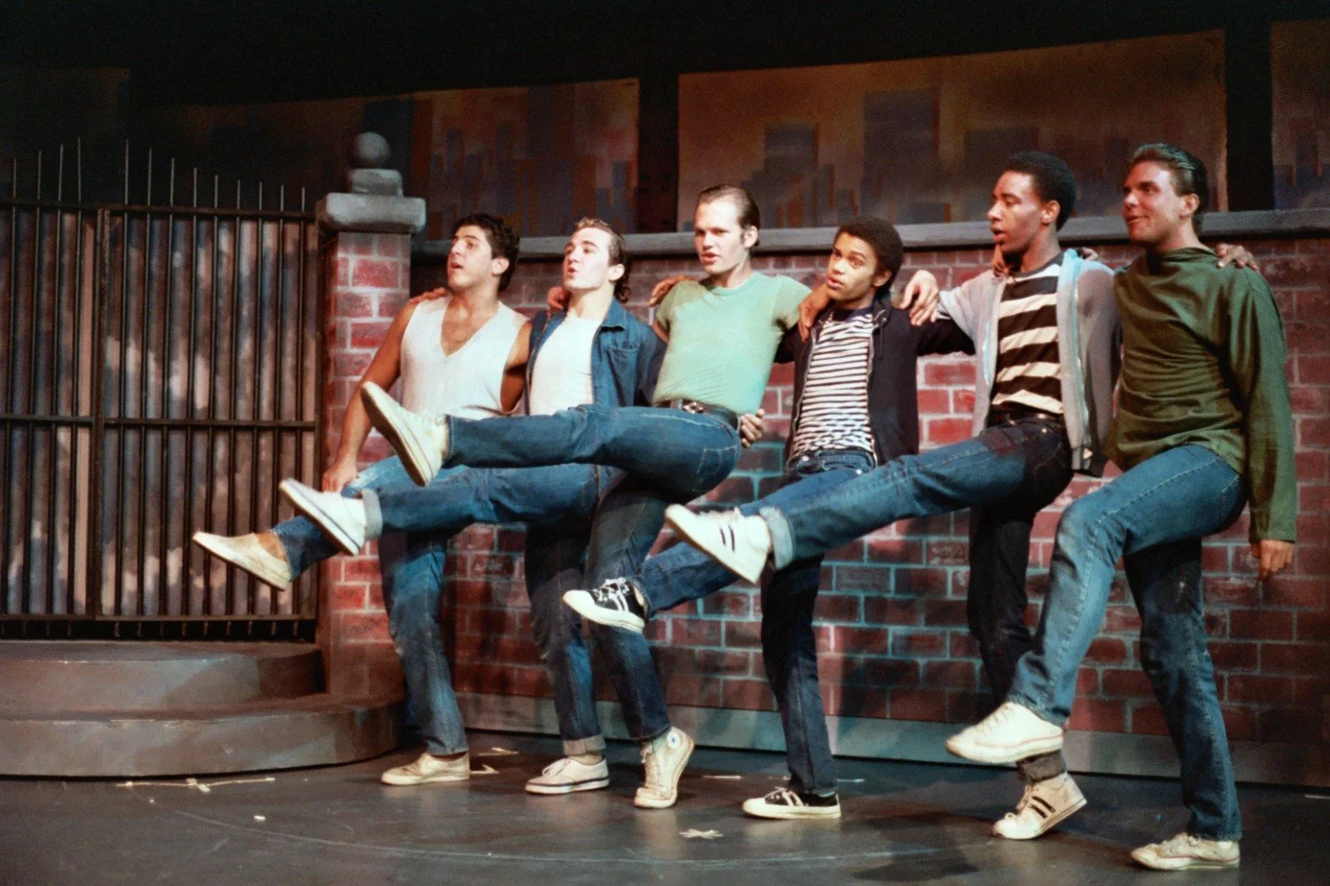 Group of six men dancing in sync with high kicks, wearing casual clothing and sneakers, in front of a brick wall set on stage.