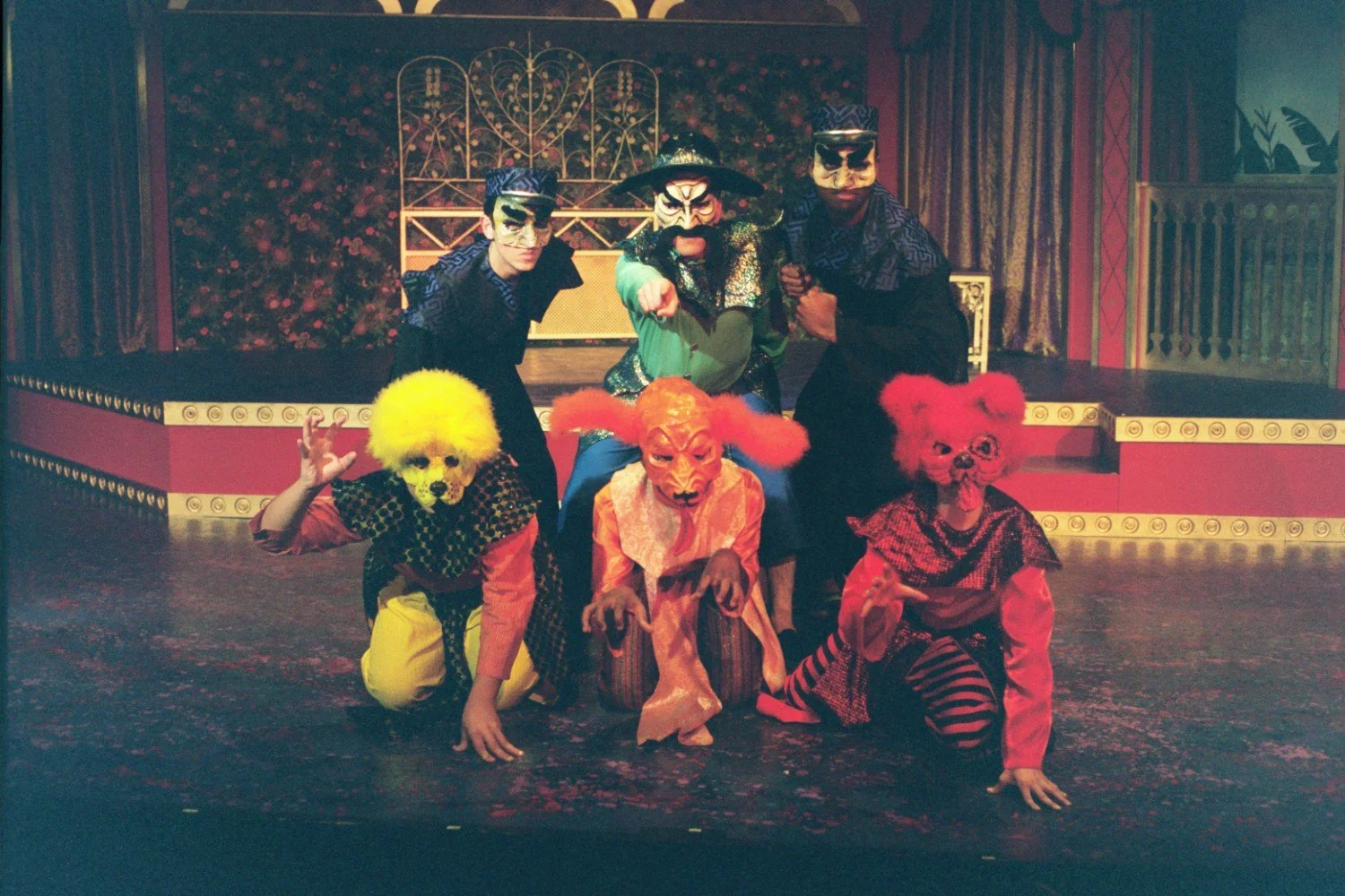 Performers in colorful costumes and masks on a stage, depicting a theatrical scene with a medieval or fantasy theme.