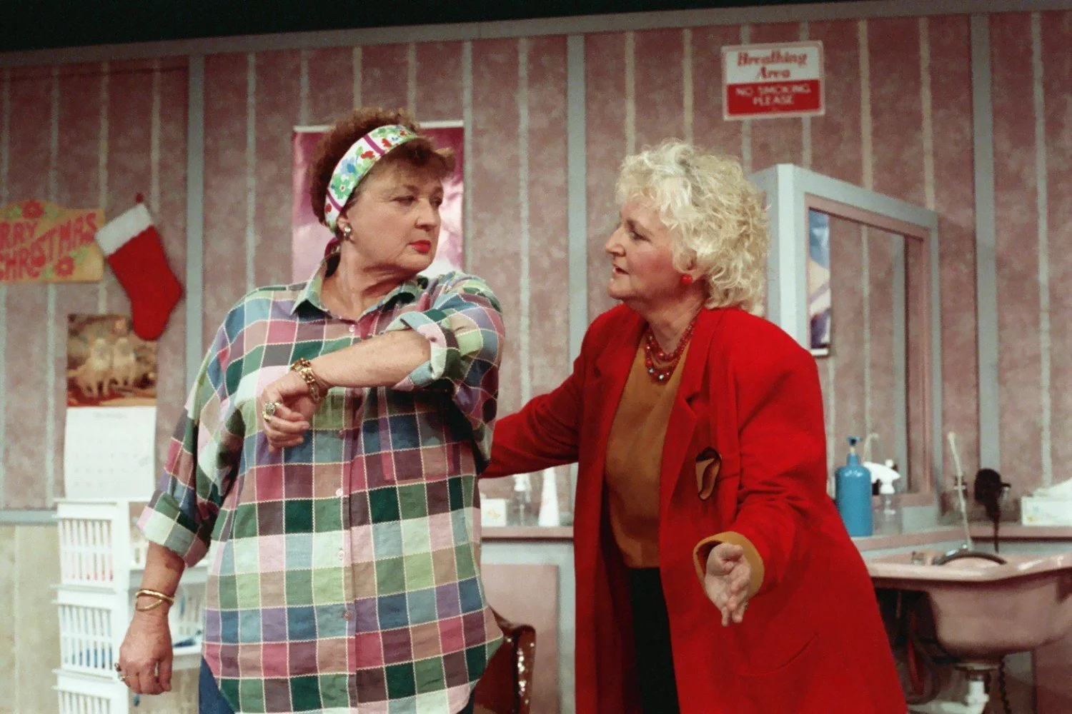 Two women in a salon setting, one wearing a colorful plaid shirt and headband, the other in a red blazer, engaged in conversation. The background has Christmas decorations and a "No Smoking" sign.