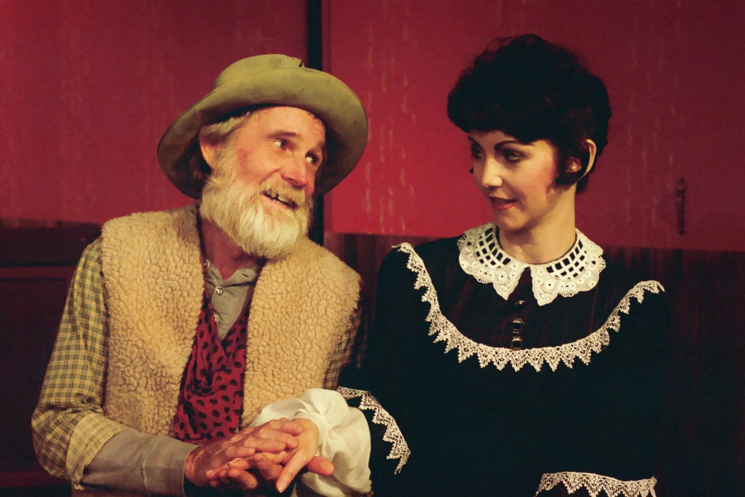 Two actors on stage, one dressed as an older man with a beard and hat, the other as a younger woman in a vintage dress, sitting closely and interacting.
