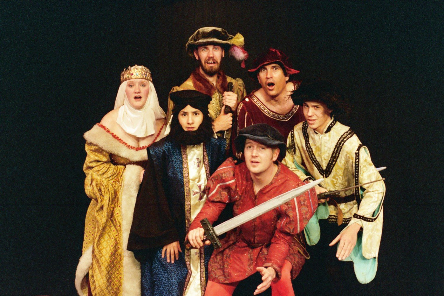 Group of actors in medieval costumes, including a person with a crown and robe, others in tunics and hats, holding a sword.
