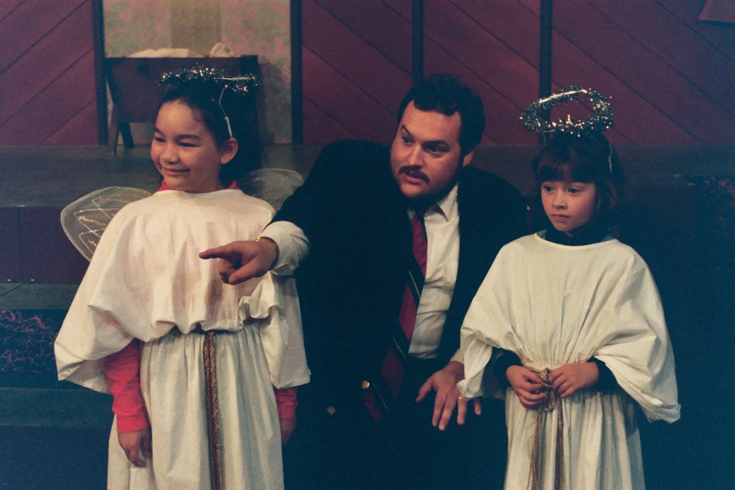 A man in a suit kneels beside two children dressed as angels, with halos and robes, on a stage setting.
