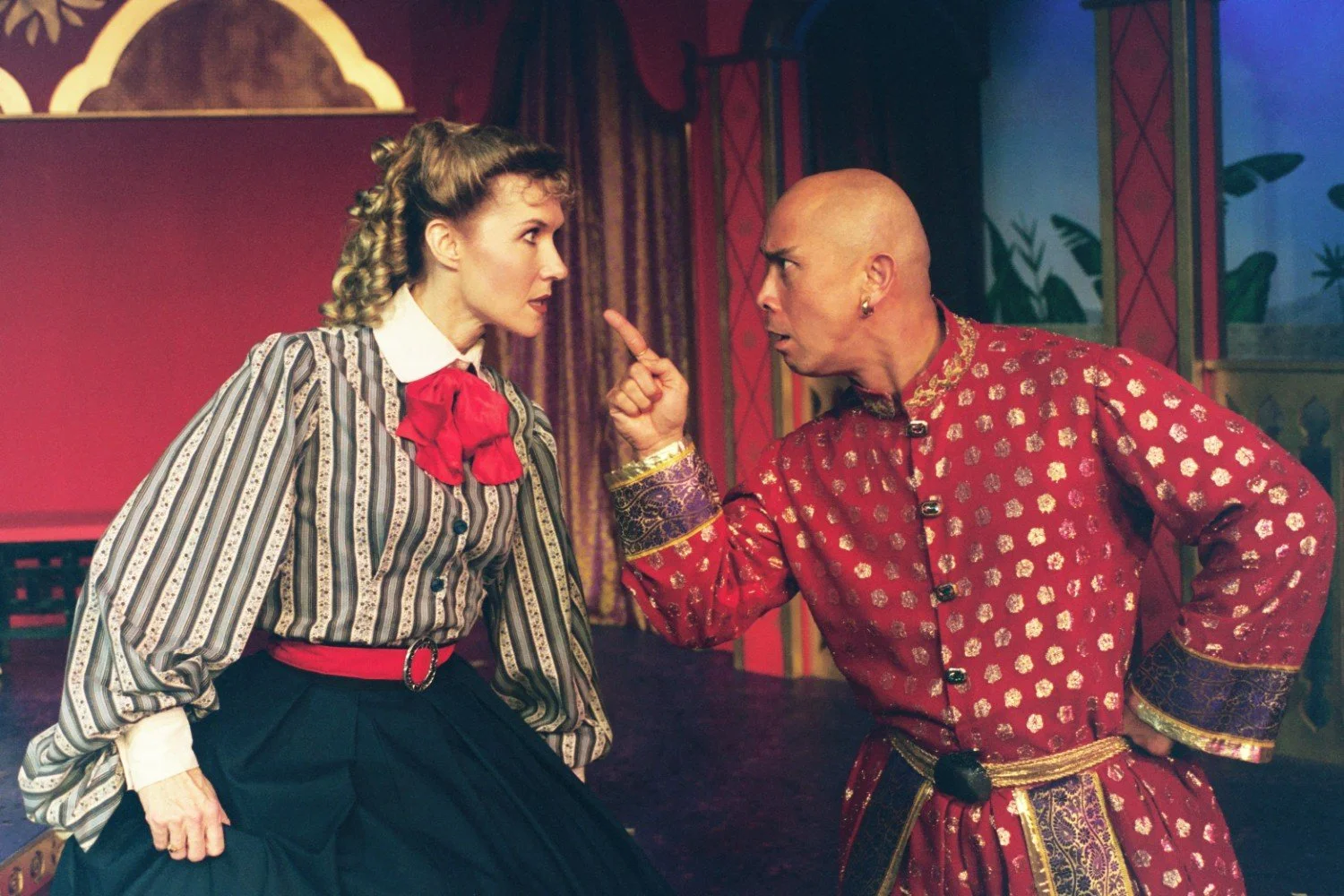 Two actors in a theatrical scene, dressed in period costumes. The woman wears a striped blouse with a red bow and a long skirt, while the man is in a red and gold embellished robe. They are engaged in a dramatic confrontation with intense expressions
