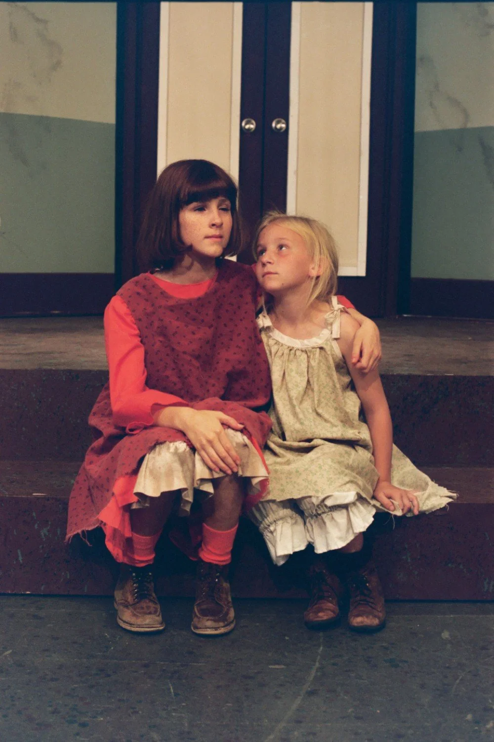 Two children dressed in vintage-style clothing sit on steps indoors, with one girl resting her arm around the other.