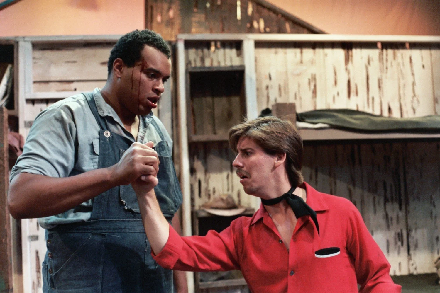Two men in a dramatic scene; one in overalls and the other in a red shirt; engaged in a hand-clasping struggle; rustic setting in the background.