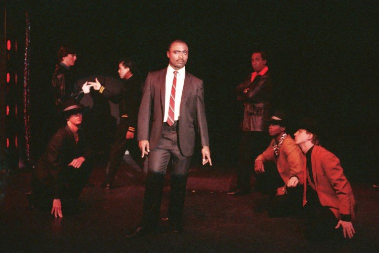 Group of performers in a theater production, with one person in a suit at the center and others in dynamic poses around the stage.