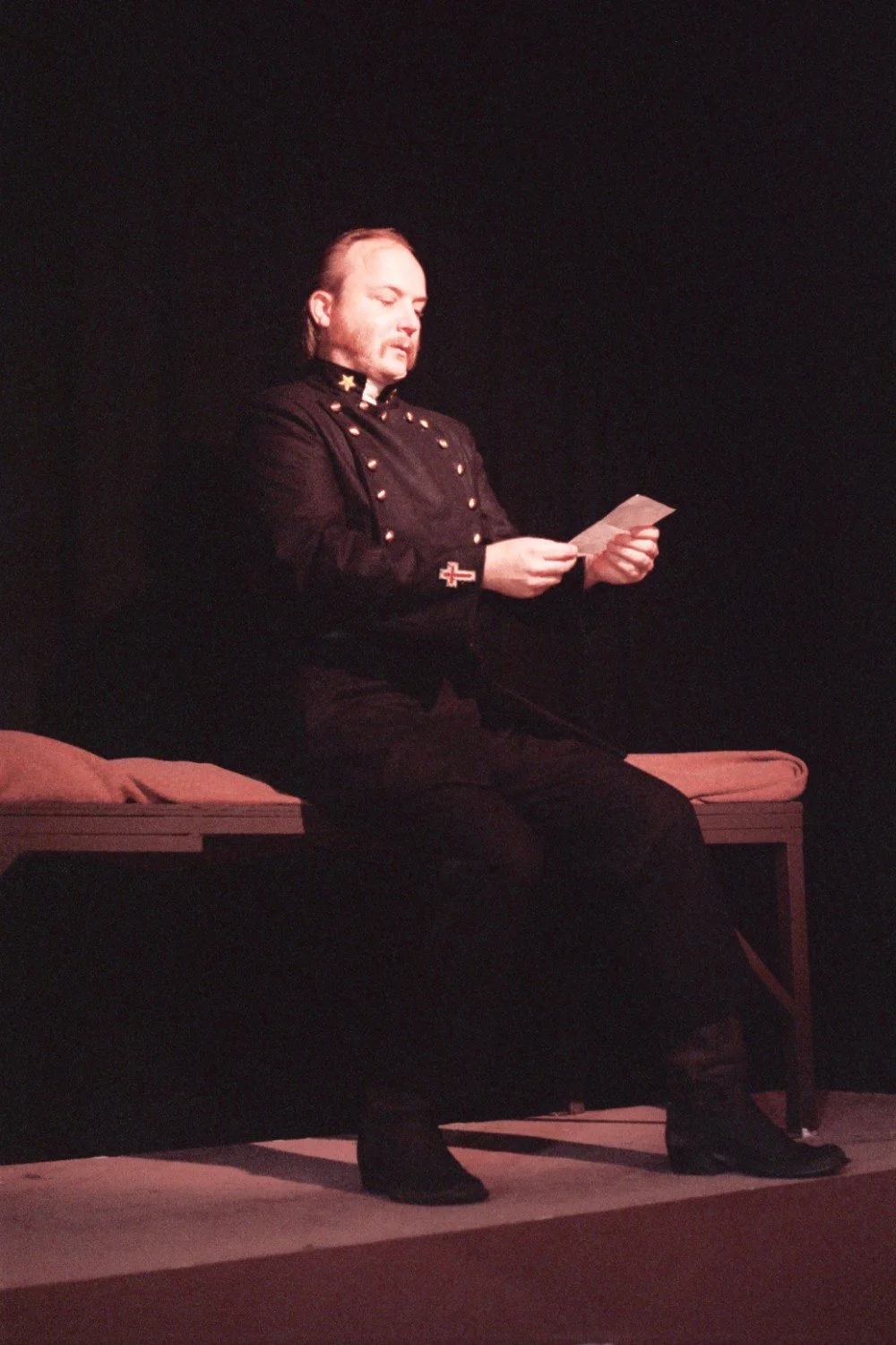 Person in military-style uniform sitting on a bench, reading a paper on stage