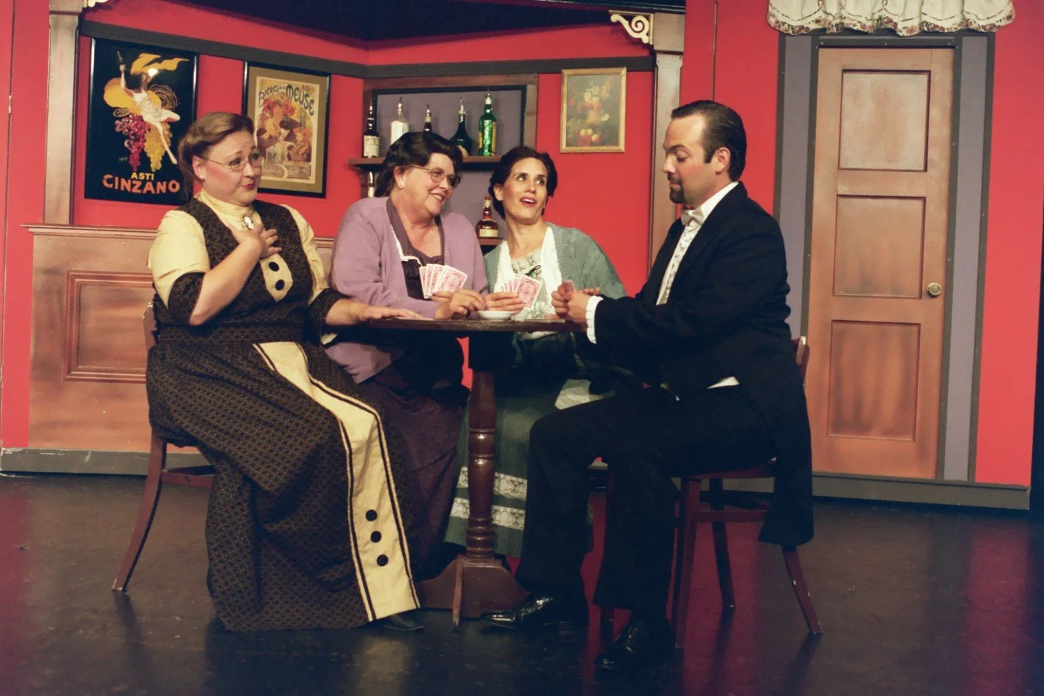 A group of four people staged in a theatrical setting, sitting around a table. Three women in period costumes hold playing cards, and a man in a suit appears to be interacting with them. The background features vintage posters and a wooden door.