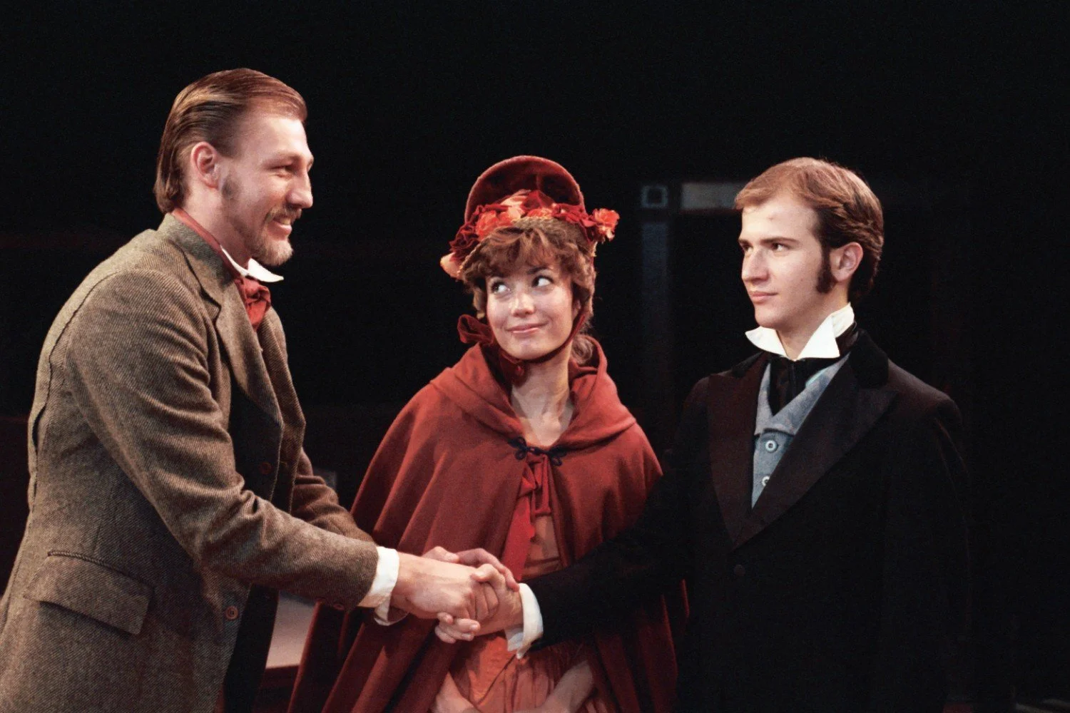 Three actors in period costumes, with two men shaking hands. A woman in a red cape and bonnet is standing between them, looking at them.
