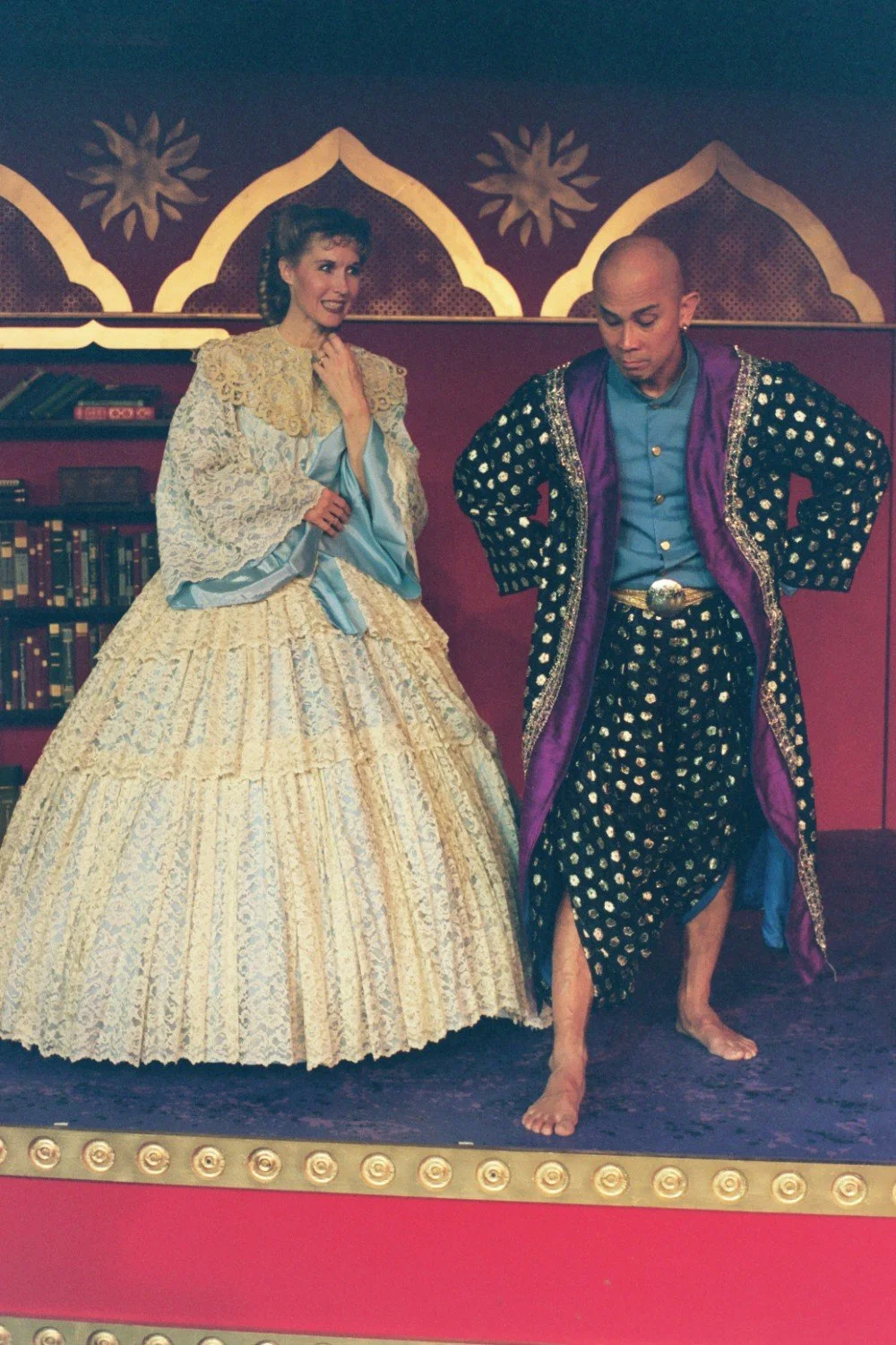A man and woman in theatrical costumes on stage, with the woman wearing a Victorian-style ball gown and the man in an ornate robe. Both are in front of a decorative backdrop.