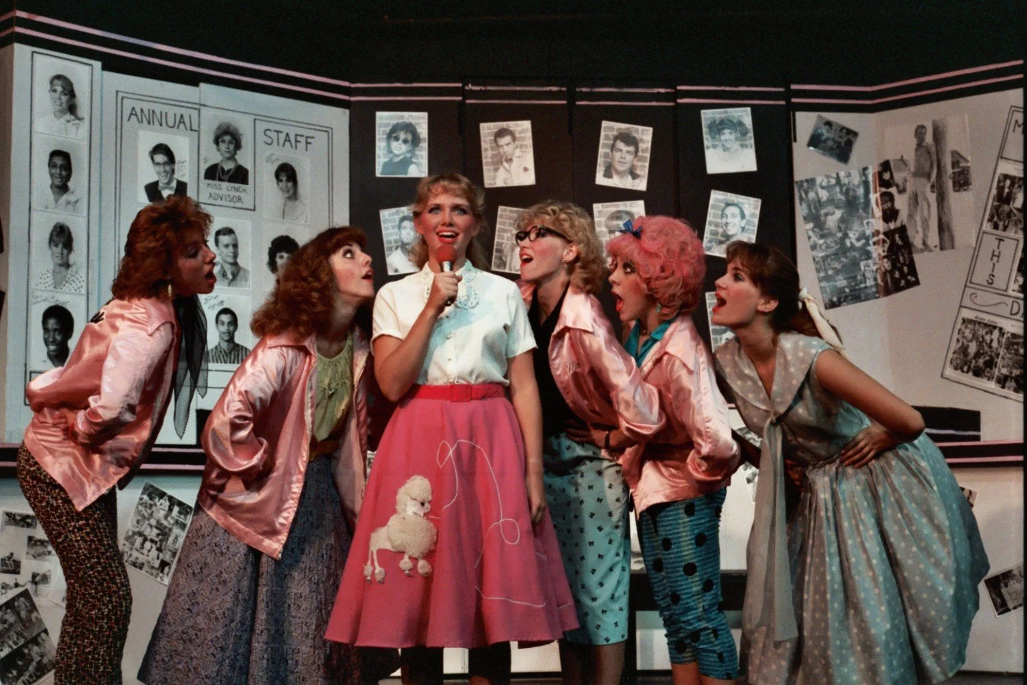 Scene from a musical featuring women in 1950s style clothing, including pink jackets and colorful skirts, one holding a microphone with others gathered around.