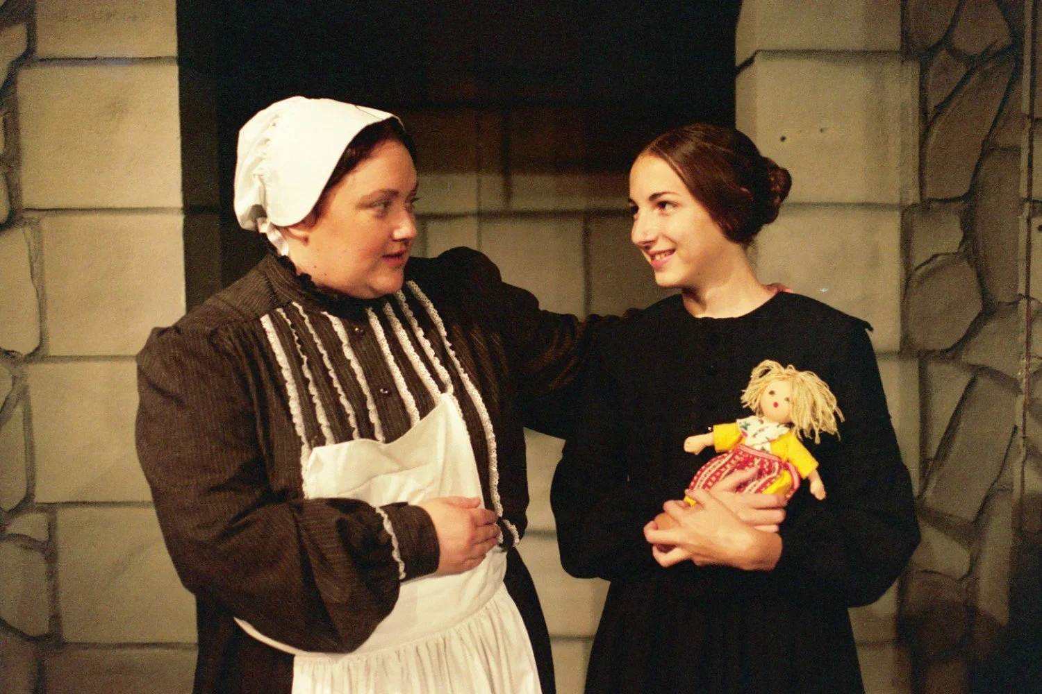 Two women in period costumes, one wearing a bonnet and apron, the other holding a doll, standing in front of a stone wall background.