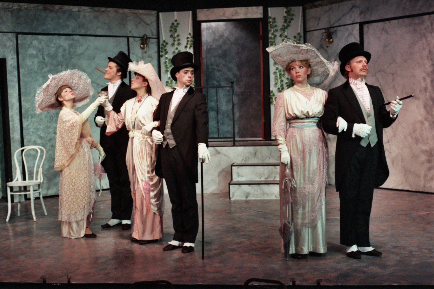 A theatrical performance featuring five actors dressed in early 20th-century formal attire, including top hats and gowns, posing on stage with props and a backdrop resembling a garden entrance.