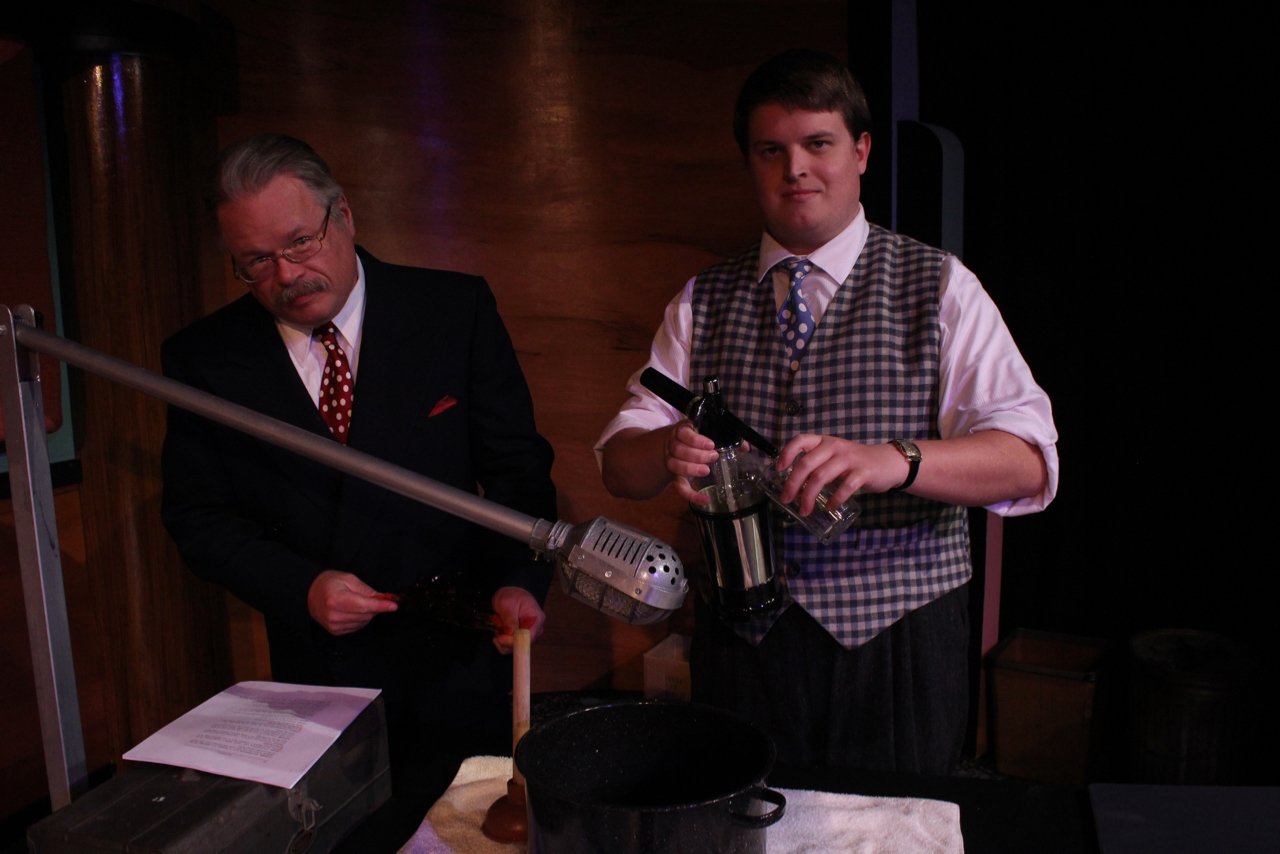 Two men performing a live sound effects demonstration using a microphone and various objects, including a bottle and a pot, on stage.
