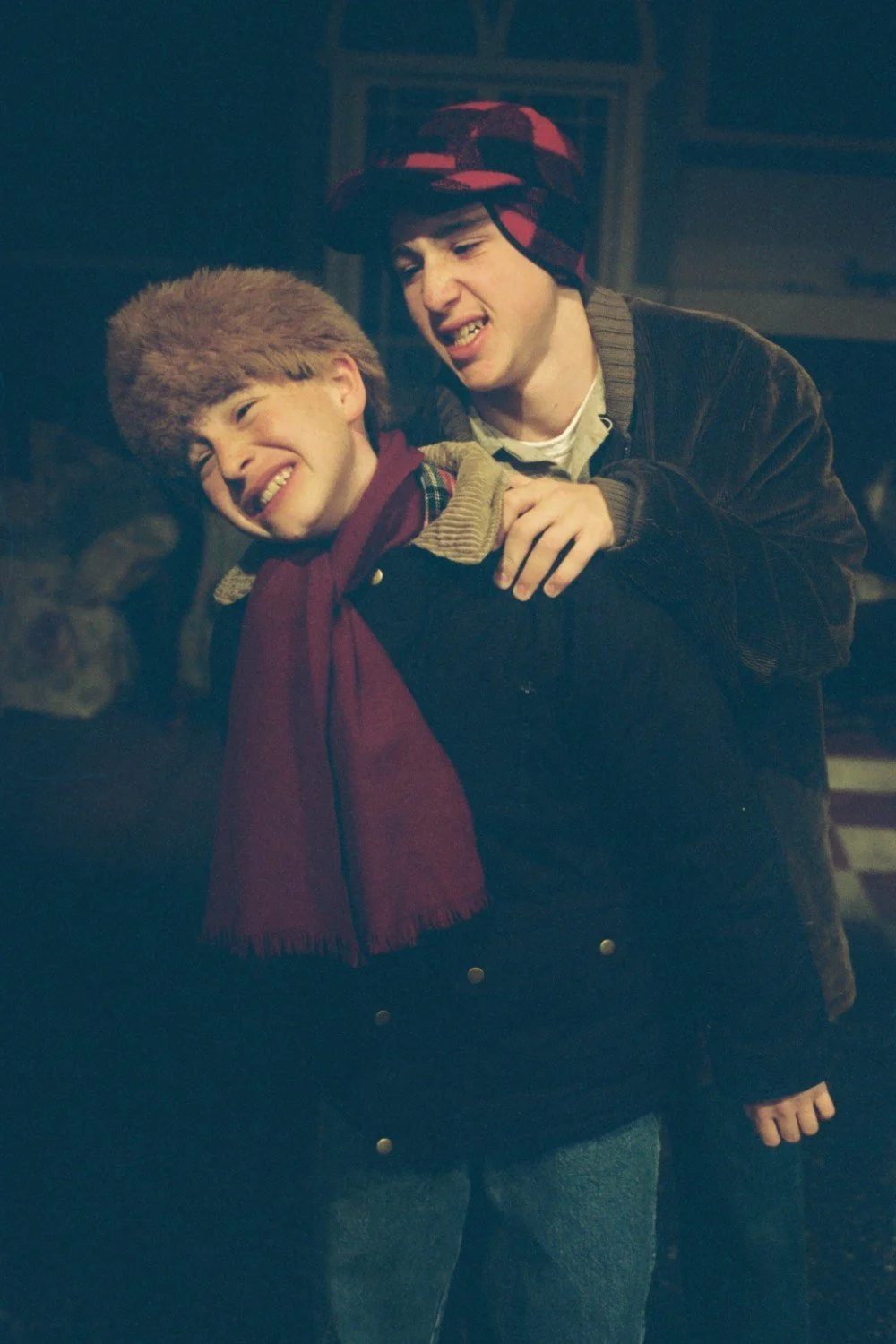 Two people dressed in winter clothing, one wearing a red scarf and fur hat, and the other in a red plaid hat, in a playful interaction indoors.
