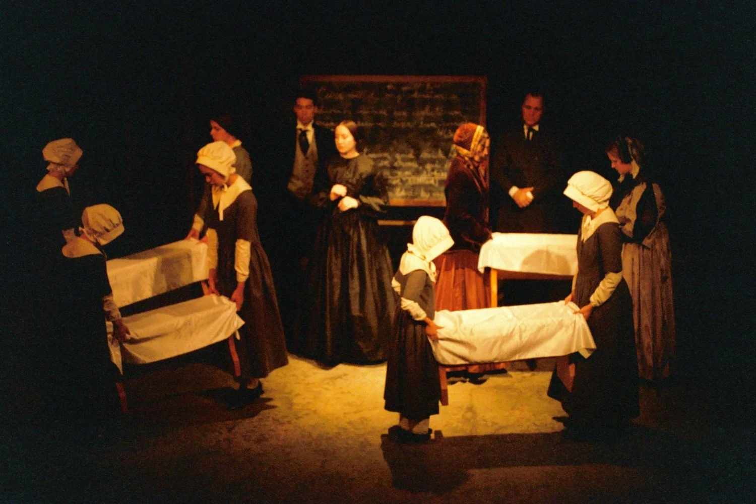 A theatrical stage scene with actors dressed in historical clothing, portraying a 19th-century setting. Several women in bonnets and dark dresses are gathered around tables covered in white cloths. A blackboard is visible in the background with two m