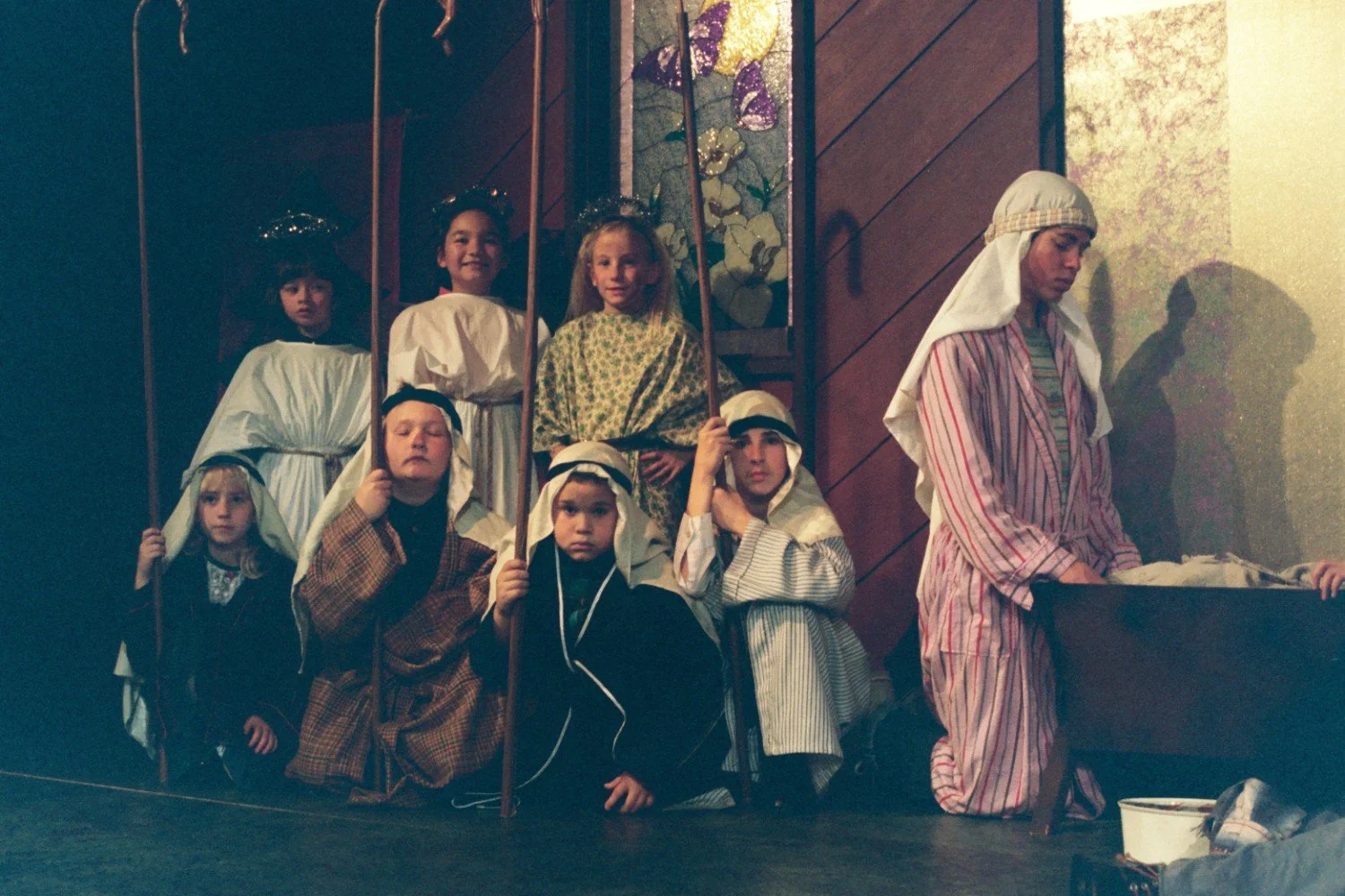 Children dressed as shepherds and angels in a nativity play on stage, with stained glass backdrop.