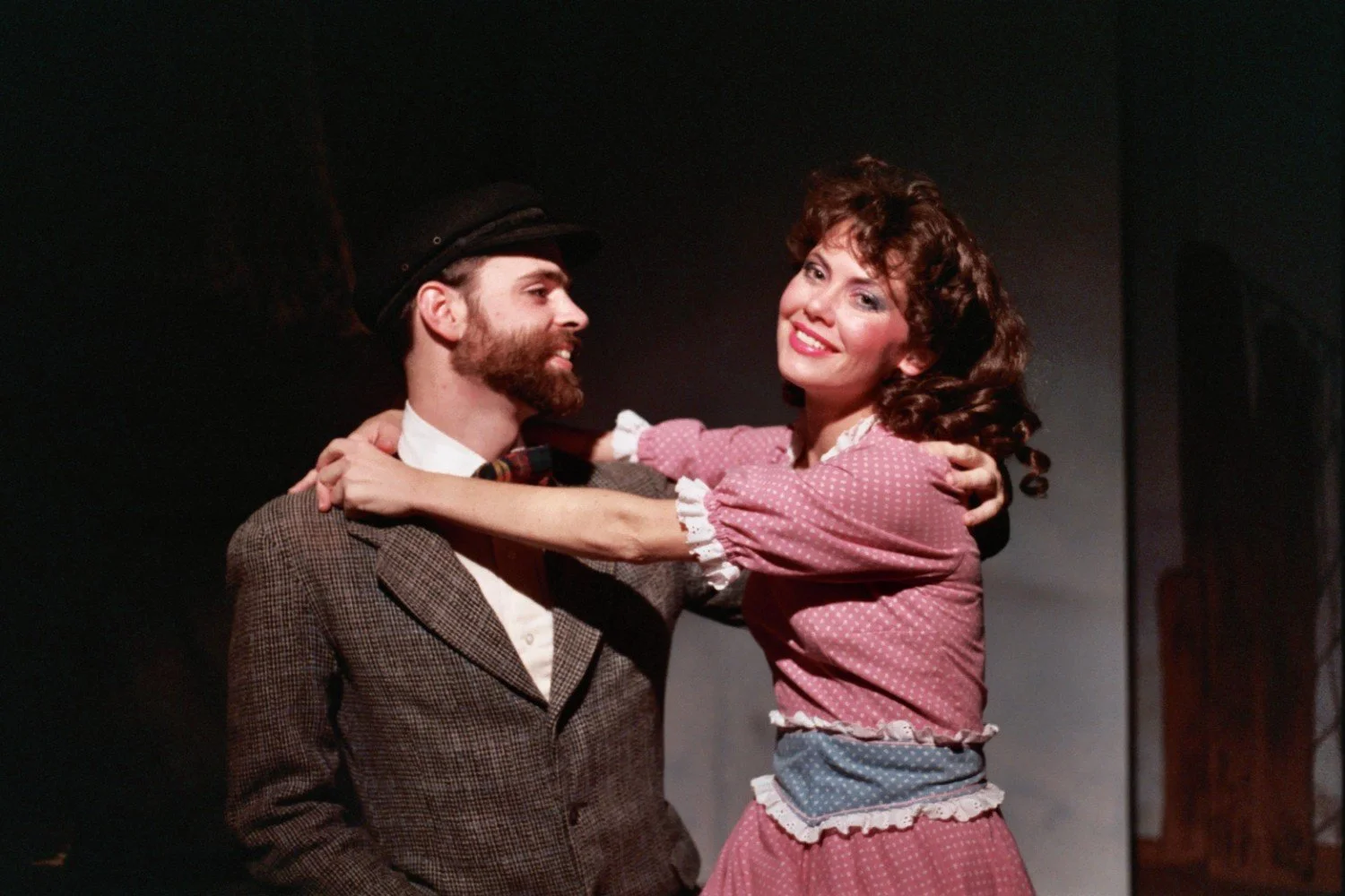 Two actors in period costumes performing on stage, with the woman smiling and holding the man's shoulders.