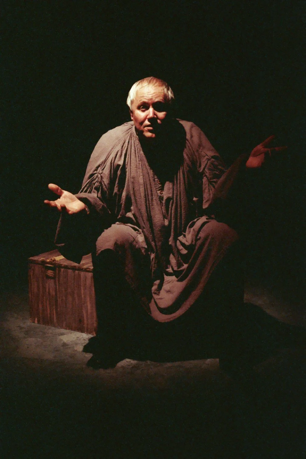 A person in dramatic lighting, wearing a robe, is seated with an expressive gesture. The setting is dimly lit, creating a theatrical atmosphere.