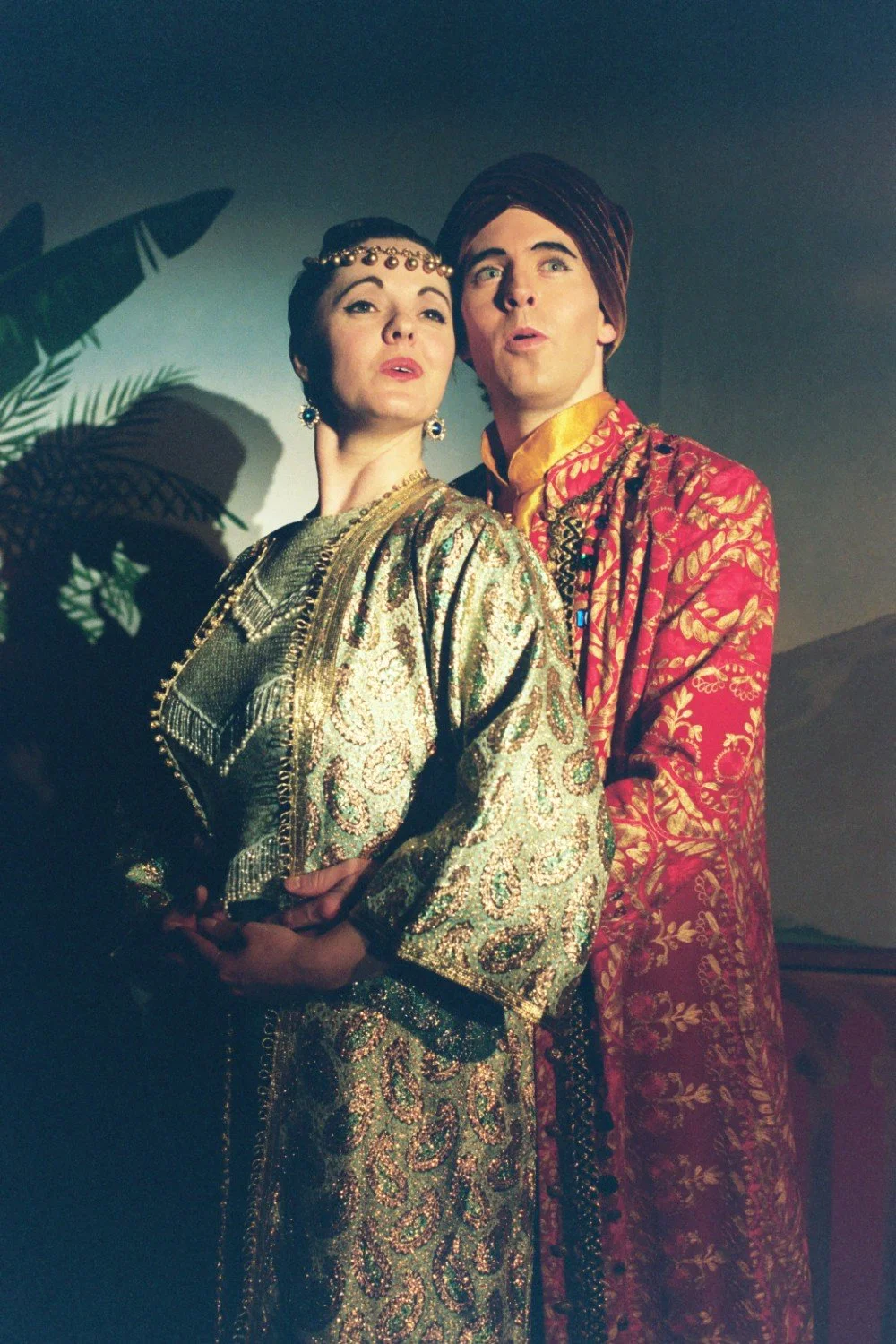 Two people in elaborate, colorful costumes perform theatrically on stage with dramatic lighting.