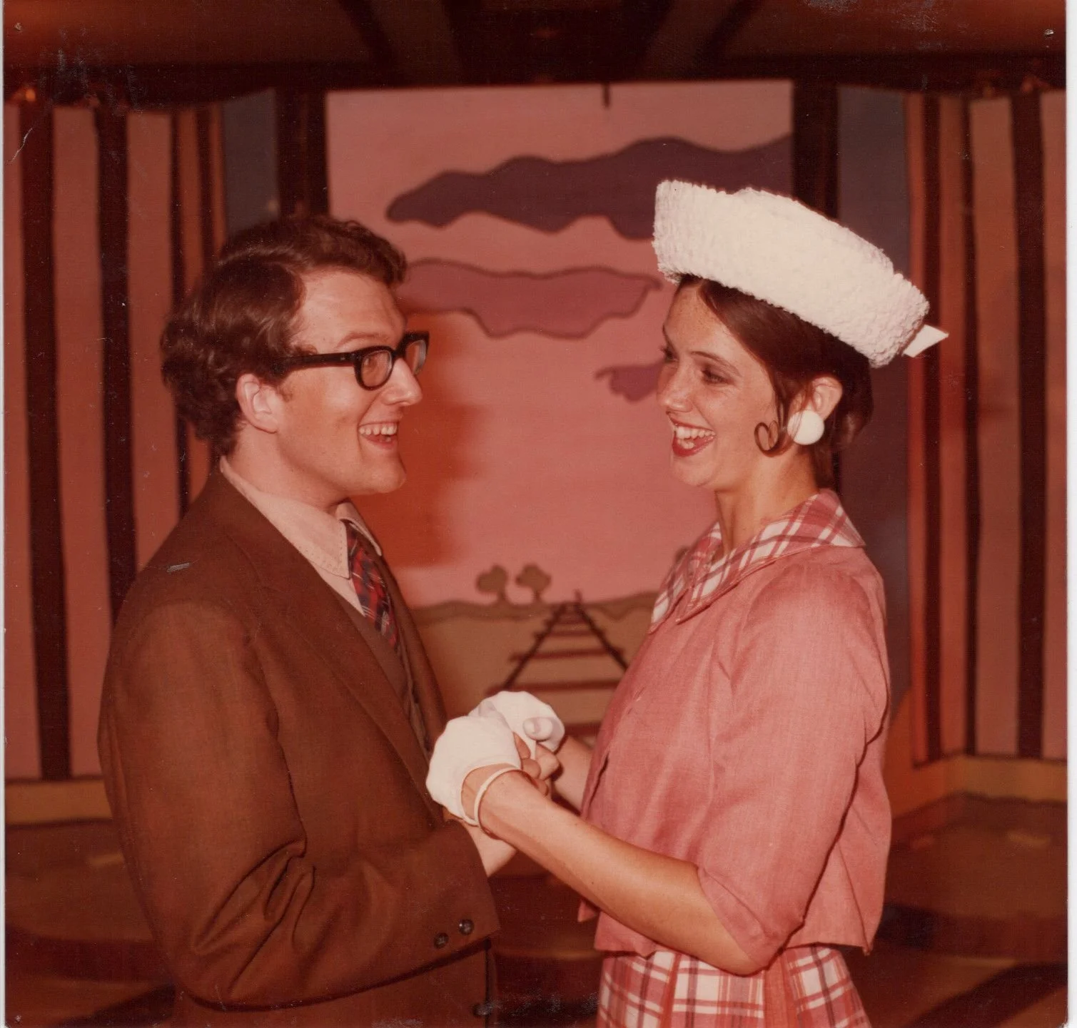 A man and a woman in vintage attire smiling at each other, with a theatrical set background.