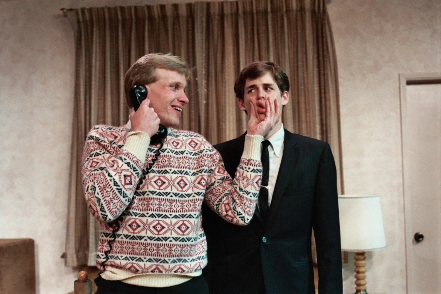 Two men indoors, one with a phone, the other wearing a suit