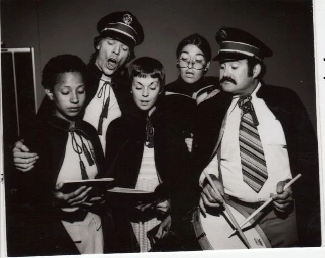 A group of five people in vintage uniforms and capes, some holding books and one holding a drum, in a black and white photo.