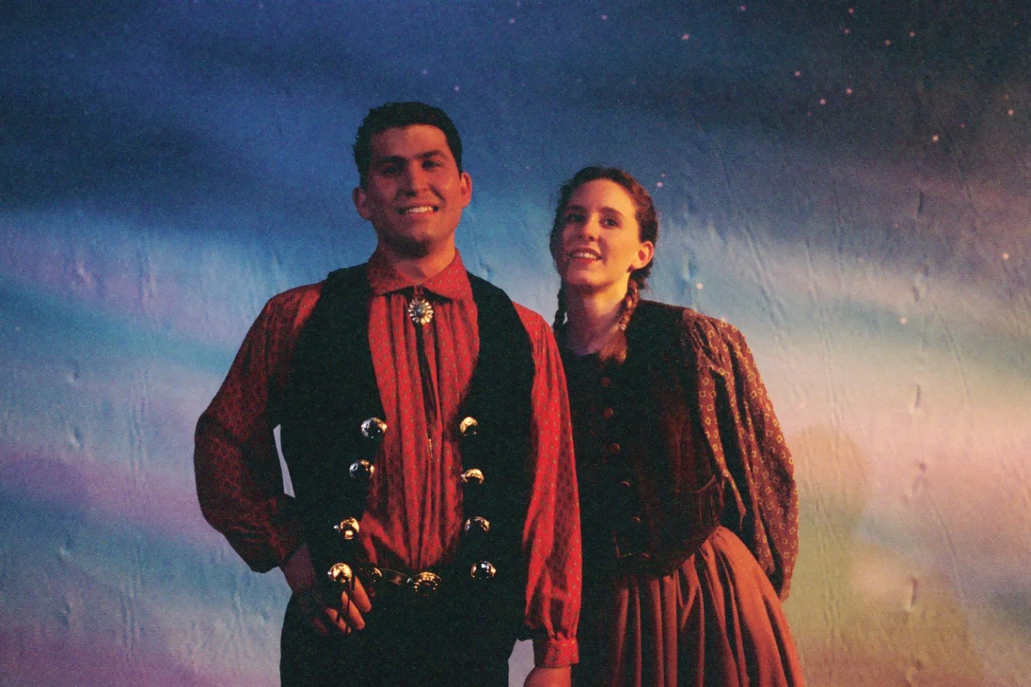 Two people in vintage or theatrical costumes posing against a colorful sky-like background.
