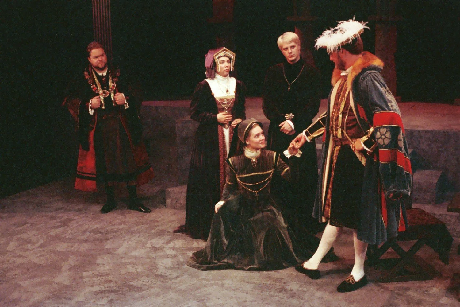 A theatrical performance featuring actors in historical costumes, possibly depicting a scene from a classic play. One actor is kneeling and receiving a gesture from another in elaborate attire, while others watch.