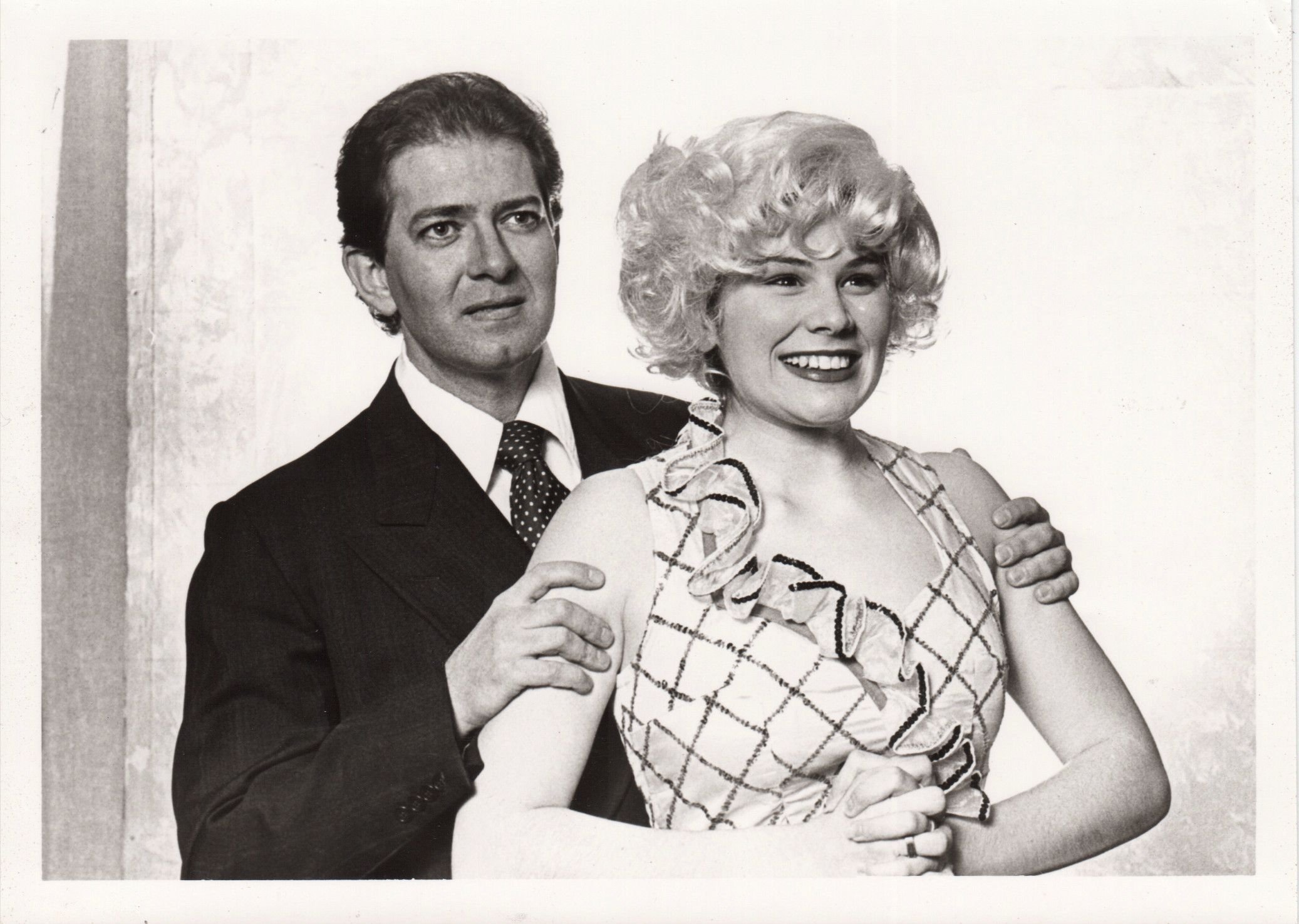 A black and white photo of a man in a suit next to a smiling woman with curly hair in a patterned dress.